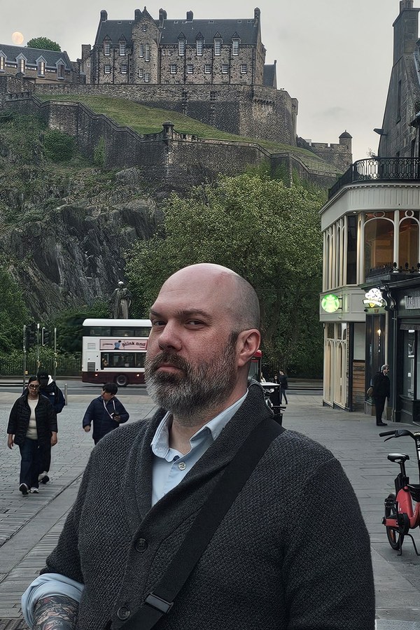 Ryan Straight, bearded and wearing a dark cardigan over a light blue shirt, stands on a street in Edinburgh in May 2026, with Edinburgh Castle on the hill behind him.